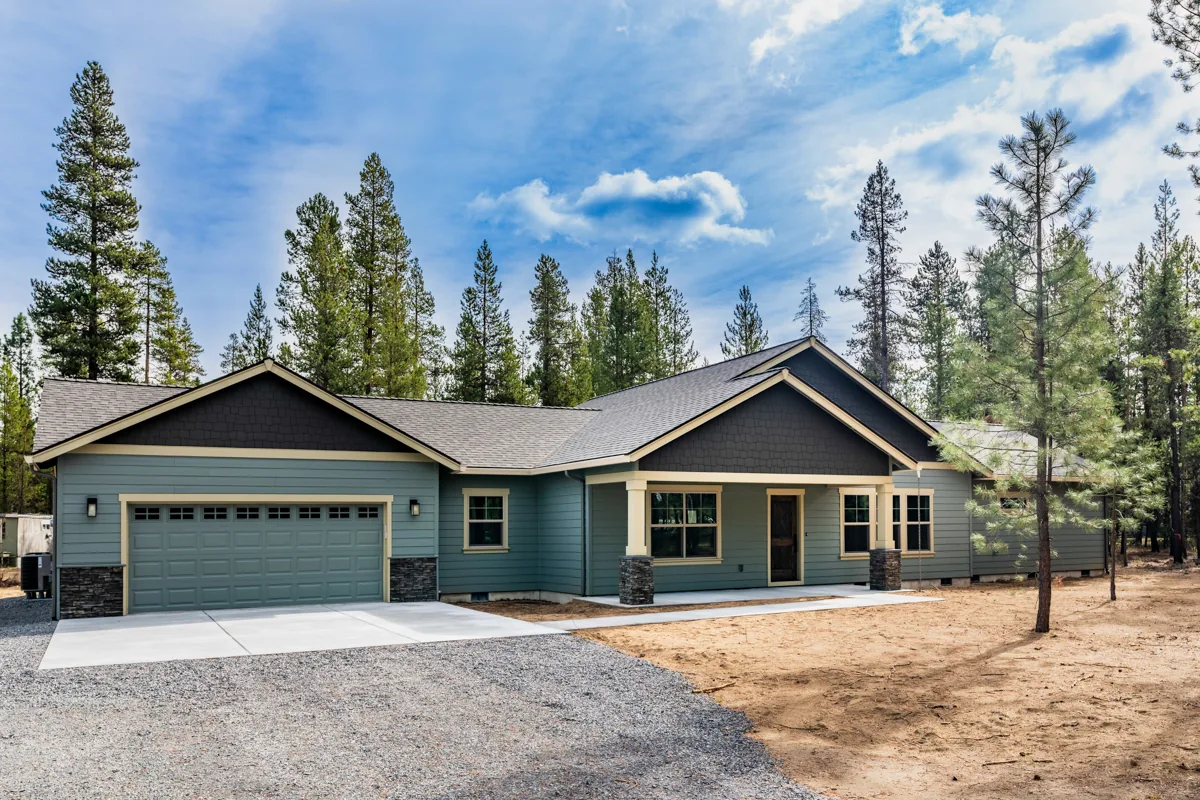 Single-story New Era Homes custom home with green siding and a stone-accented garage on a wooded Central Oregon lot